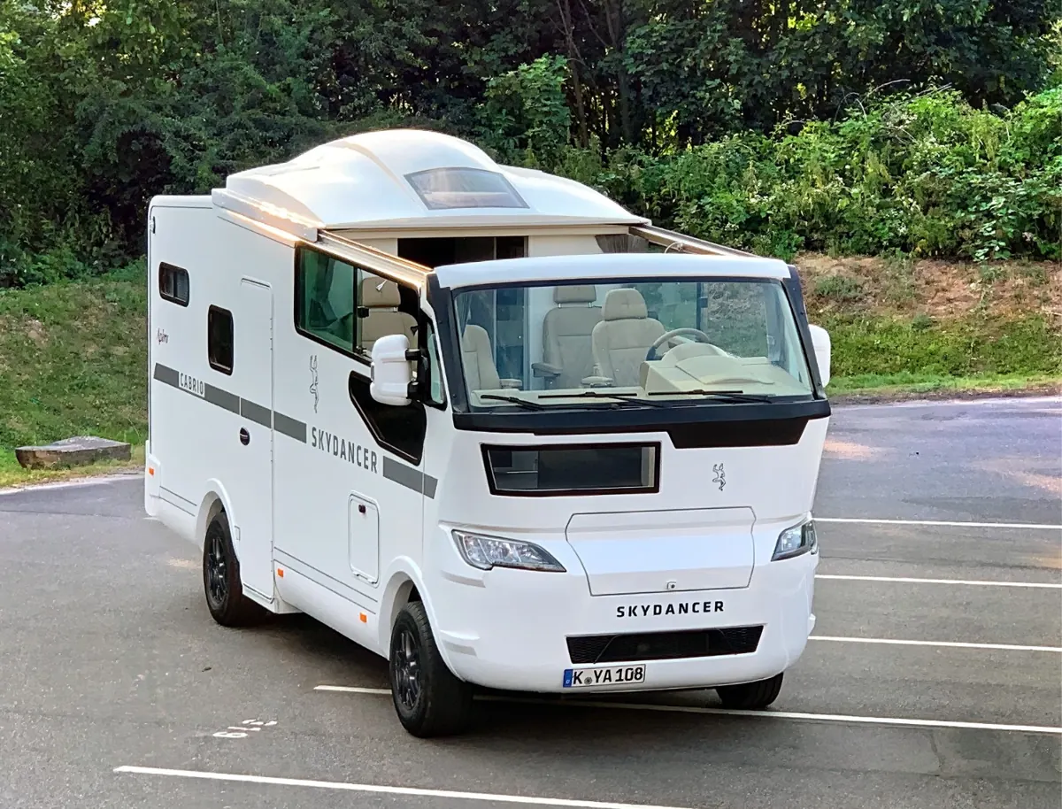 Weißes Skydancer-Reisemobil auf einem Parkplatz, das Cabrio-Dach über dem Wohnraum ist geöffnet.